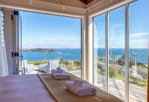 A bedroom with a large window, a bed with pillows, and two towels on a wooden platform. - The Belle House – Where Sea Meets Sky (Waiheke Island)