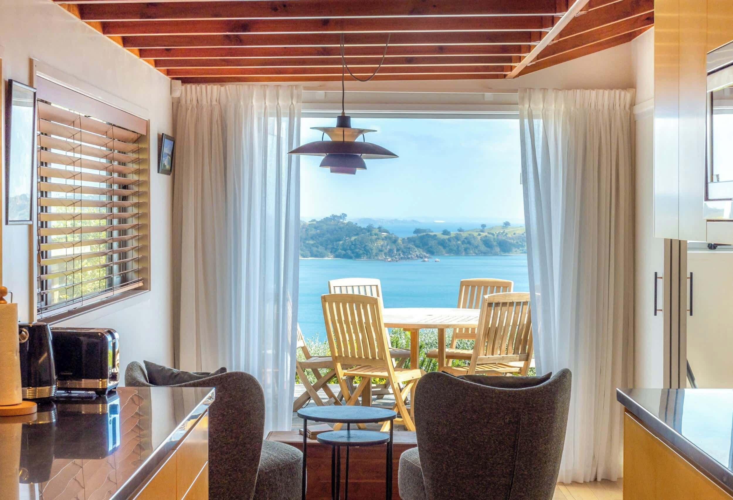 A room with a view of the sea, a table with chairs, a hanging light fixture, and a window with blinds.