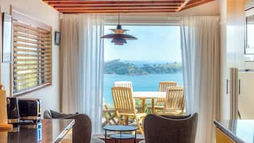 A room with a view of the sea, a table with chairs, a hanging light fixture, and a window with blinds.