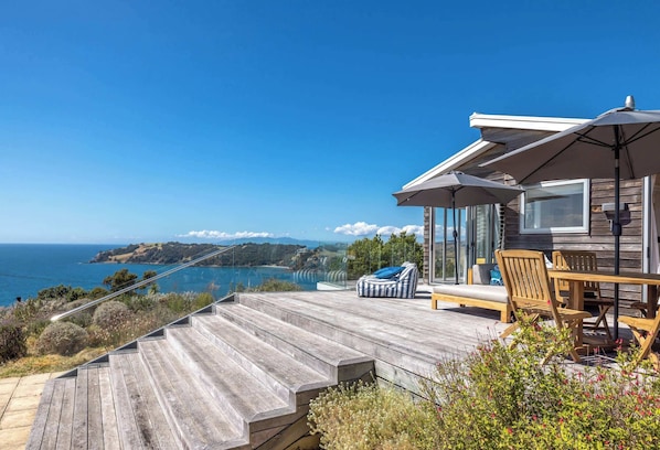 A wooden deck with outdoor furniture, a glass balustrade, and a view of the ocean. - The Belle House – Where Sea Meets Sky (Waiheke Island)