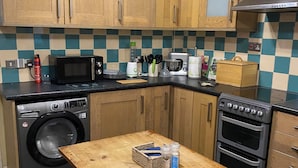 A kitchen with wooden cabinets, a washing machine, a microwave, a toaster, and a table with a basket and bottles. - Cosy Cottage style Bungalow Close to Leicester City, Abbey Park & The Highcross (Leicester)