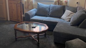 A living room with a sectional sofa, a glass coffee table, a wooden door, and a window with floral curtains. - Cosy Cottage style Bungalow Close to Leicester City, Abbey Park & The Highcross (Leicester)