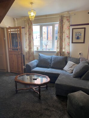 A living room with a sectional sofa, a glass coffee table, a wooden door, and a window with floral curtains. - Cosy Cottage style Bungalow Close to Leicester City, Abbey Park & The Highcross (Leicester)