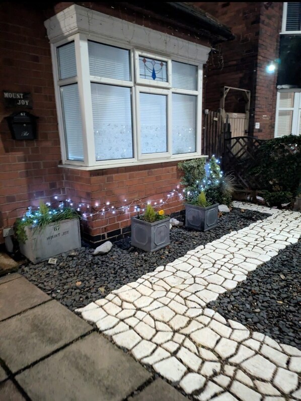 A brick building with a white window, a stone pathway, and potted plants with lights.