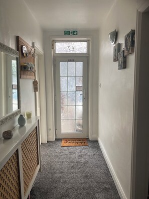 A hallway with a frosted glass door, a fire exit sign, a welcome mat, and a mirror with decorative items. - Cosy Cottage style Bungalow Close to Leicester City, Abbey Park & The Highcross (Leicester)