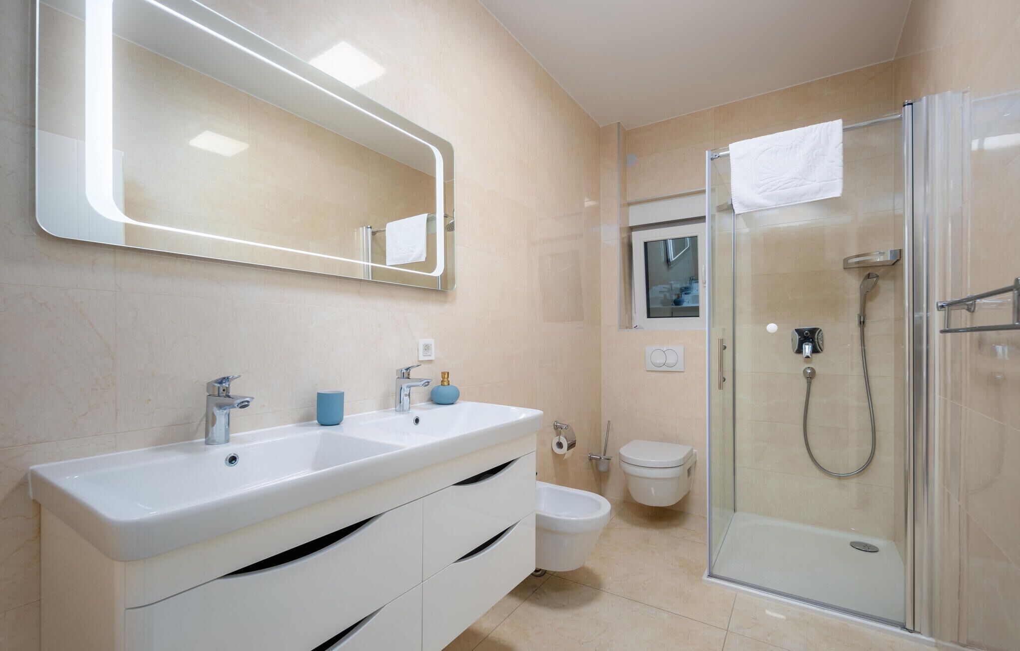 Un baño moderno con un espejo grande, dos lavamanos, inodoro y ducha con mampara de vidrio.