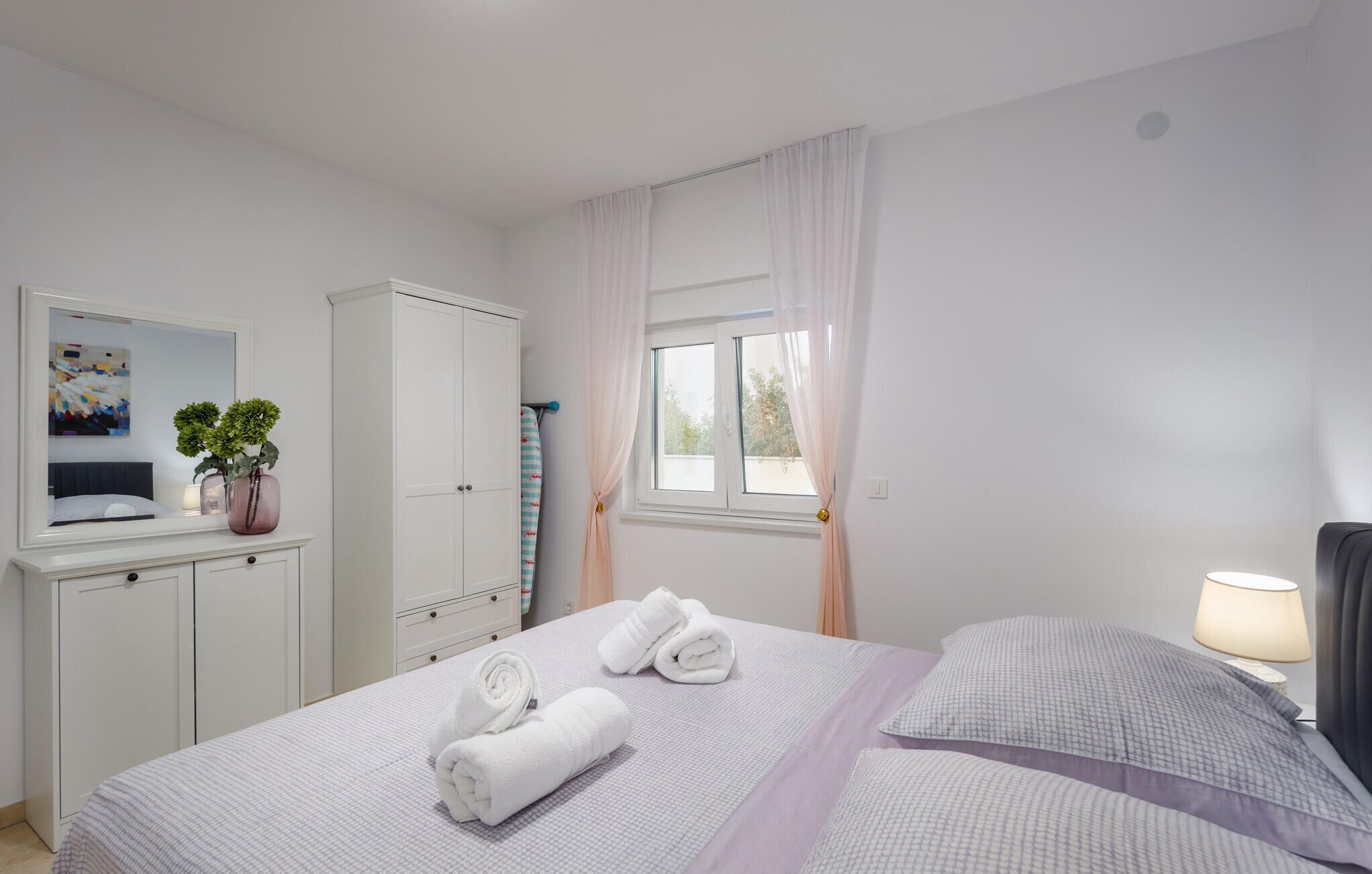 Hotellihuone, jossa on sänky, vaatekaappi, peili, ikkuna verhoilla ja yöpöytälamppu.