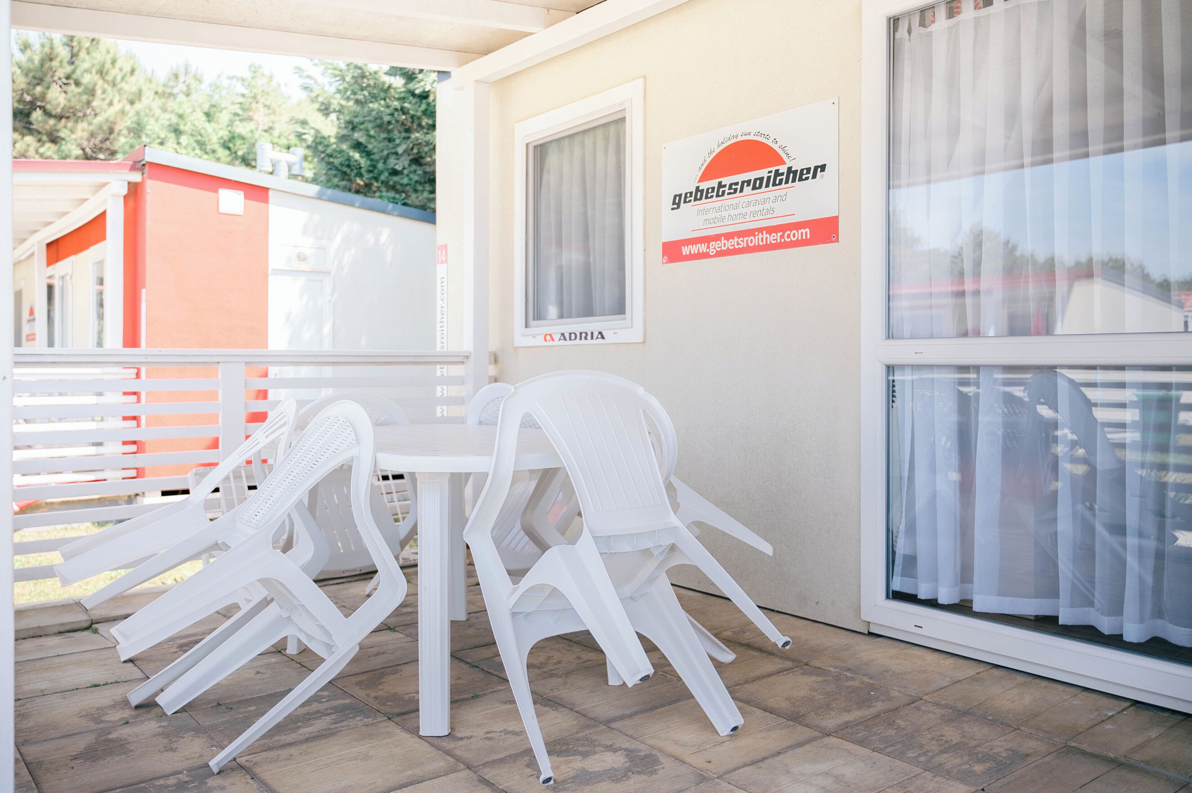 A covered outdoor area with a table and chairs, a window with curtains, and a sign for a caravan and mobile home rental service.