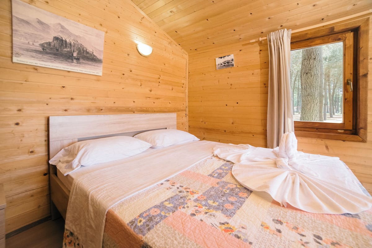 A wooden cabin bedroom with a bed, a painting on the wall, and a window with a curtain.