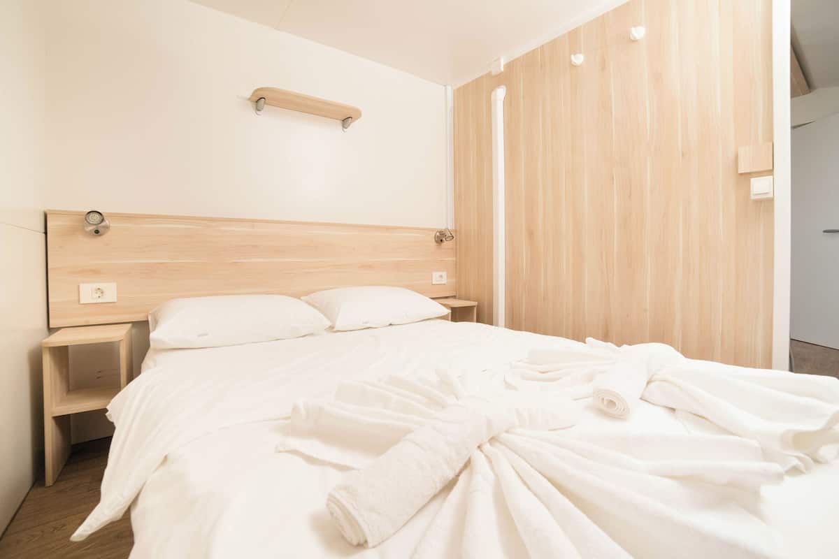 A hotel room with a wooden headboard, a bedside table, a wall-mounted shelf, and a neatly made bed with white bedding and a rolled towel.