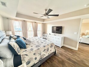 Une chambre à coucher comprenant un lit, un ventilateur de plafond, une commode, un téléviseur et une fenêtre avec des rideaux.