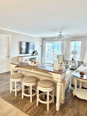 A spacious living area with a central island, bar stools, a television, and a balcony with outdoor furniture. - Welcome to Far Far Away at Ocean Marsh! (North Myrtle Beach)