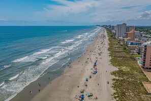 A coastal beach with a long stretch of sand, a few umbrellas, and buildings along the shore. - Welcome to Far Far Away at Ocean Marsh! (North Myrtle Beach)