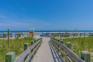 Un sentier qui longe la plage et mène à une étendue de sable où l’on trouve des parasols et des chaises de plage.