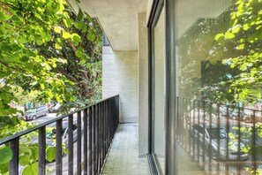 Un balcón estrecho con barandilla de metal, pared de concreto y vista a una calle con coches estacionados y áreas verdes.