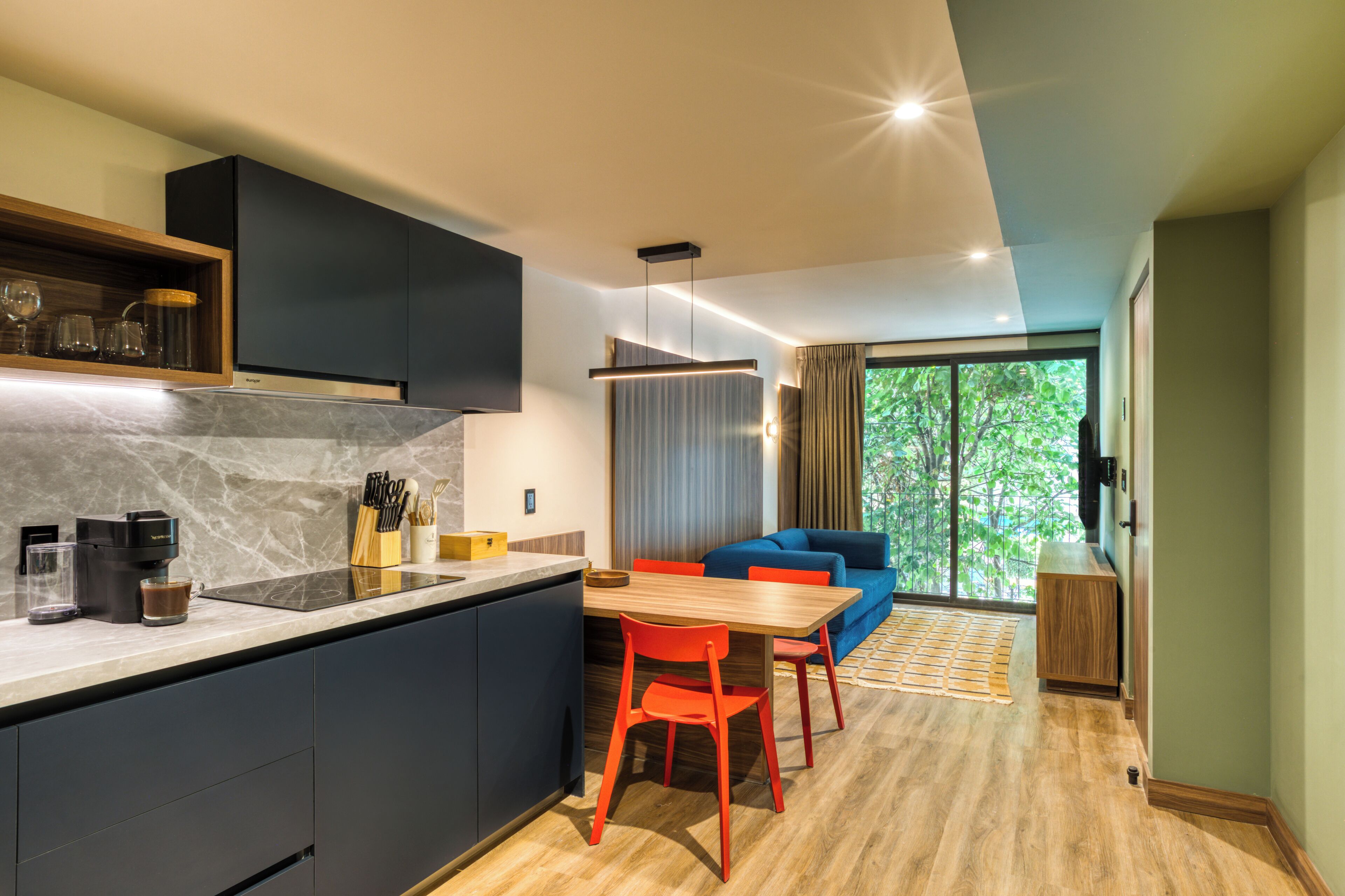 A modern kitchen with dark cabinets, a marble countertop, and a dining area with a wooden table and red chairs.