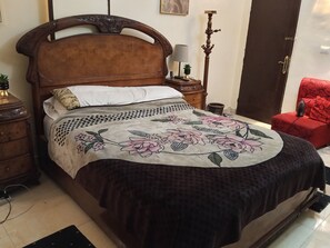 Ein Schlafzimmer mit einem hölzernen Bett, einem roten Sessel, einem Nachttisch, einer Lampe und einem an der Wand befestigten Regal.