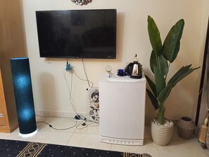 Ein Zimmer mit flachbildfernseher an der Wand, einem weißen Kühlschrank, einem blauen zylinderförmigen Lautsprecher, einer Zimmerpflanze und einem Wanddekor.