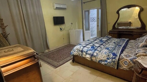 Ein Schlafzimmer mit einer Chandelüster, einem Bett mit gemusterten Betttextilien, einem Holzschrank, einem Spiegel und einer Heizung.