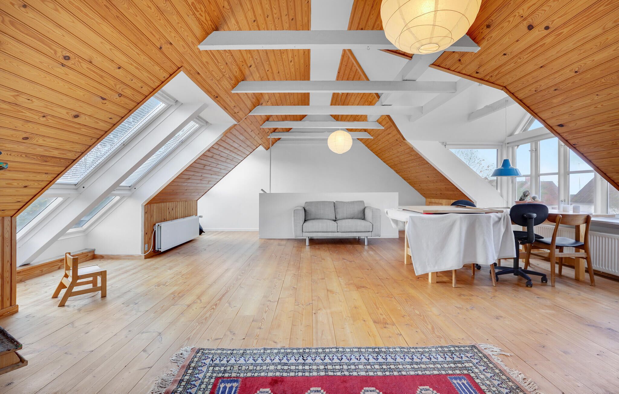 A spacious attic room with wooden beams, a grey sofa, a dining table, and a large window.