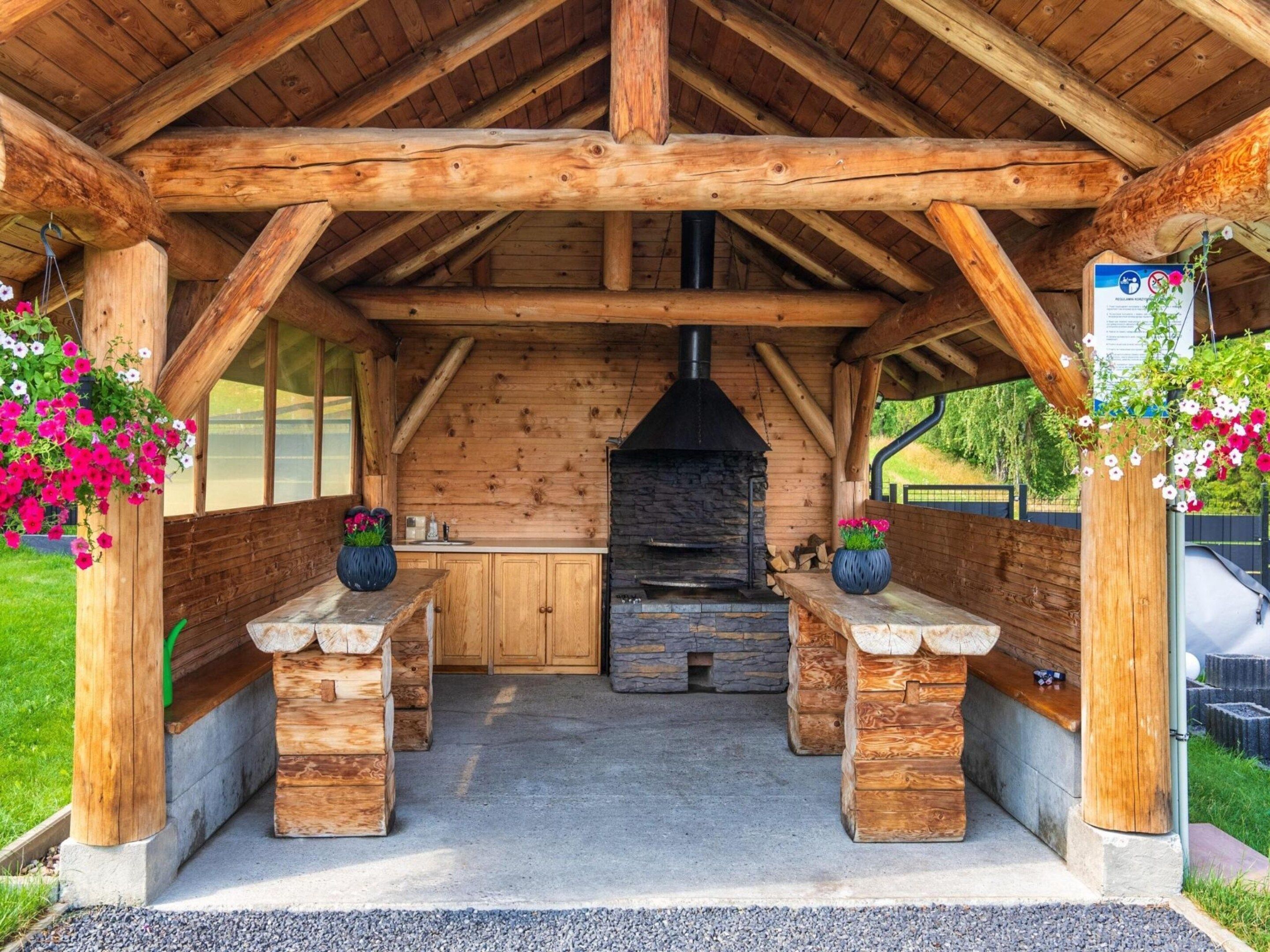 A wooden outdoor kitchen area with a stone fireplace, wooden benches, and flower baskets.
