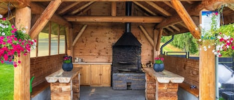 A wooden outdoor kitchen area with a stone fireplace, wooden benches, and flower baskets.