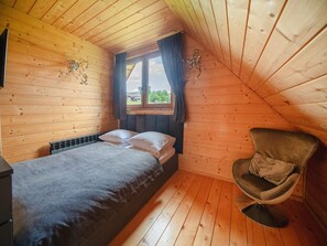 A small, cozy room with wooden walls and a sloped ceiling, featuring a bed, a chair, and a window with curtains. - Luxury Holiday Villa, Pool, Sauna, Jacuzzi (Nowy Targ)