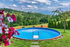 Ein runder Pool mit einer Leiter, umgeben von einer Holzterrasse und Rasen, mit bewaldeter Landschaft im Hintergrund.