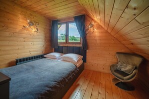 Una habitación de cabaña de madera con una cama, una silla, una ventana con cortinas y una lámpara fijada a la pared.