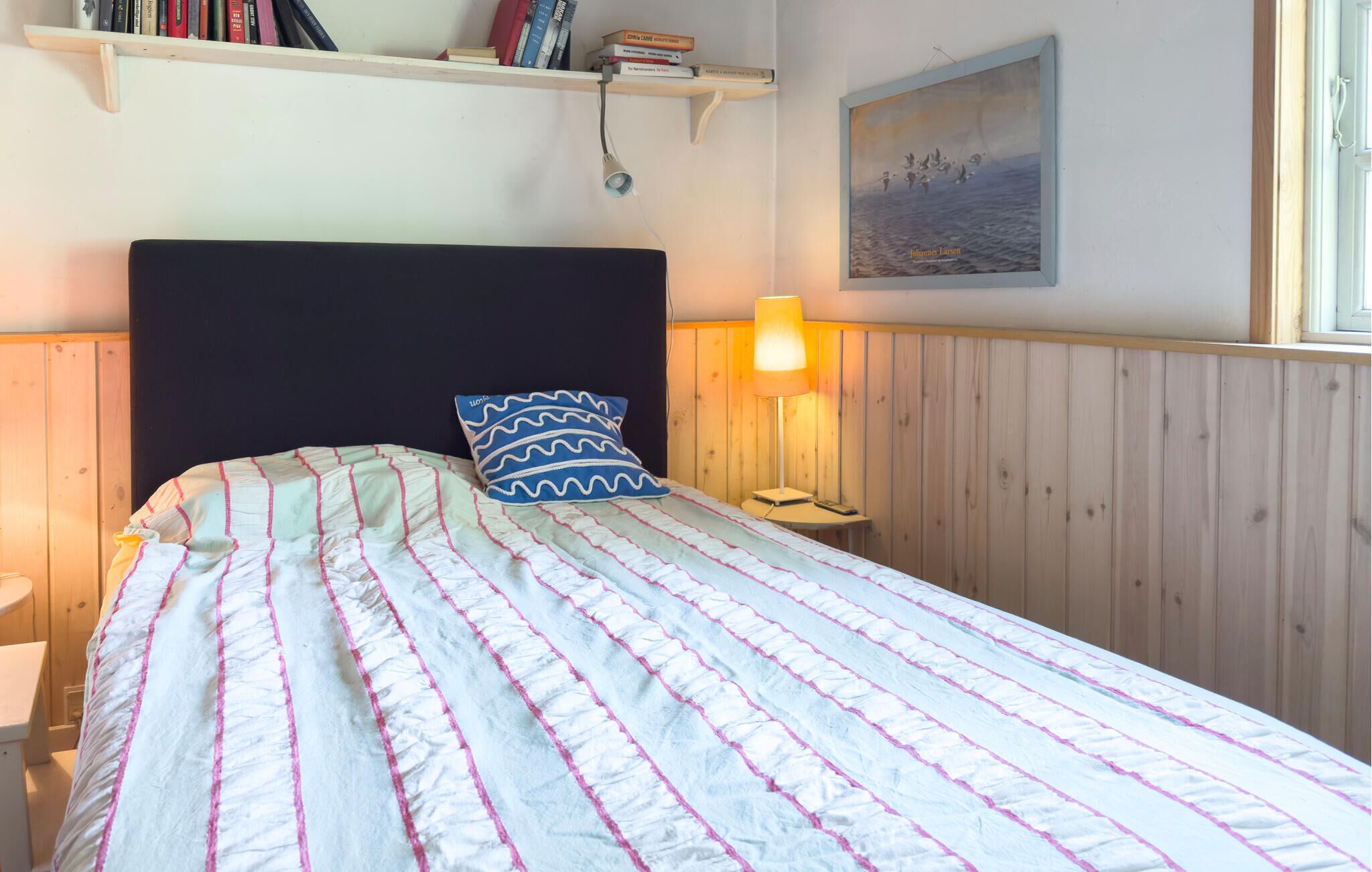 Ein Bett mit gestreifter Decke, eine Nachttischlampe, ein Nachttisch und ein Regal mit Büchern.