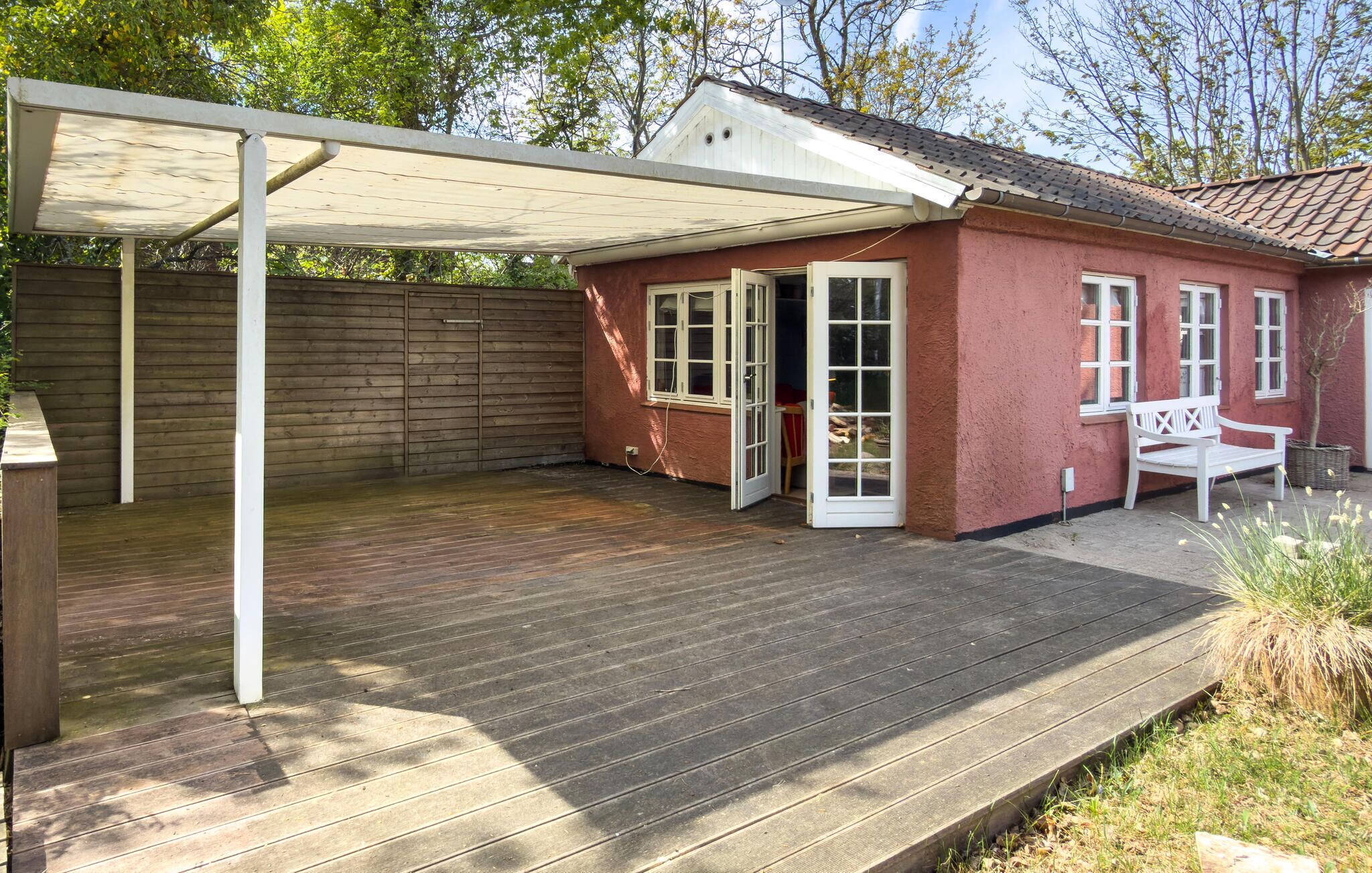 A covered patio area with a wooden deck, a white bench, and a red building with windows.