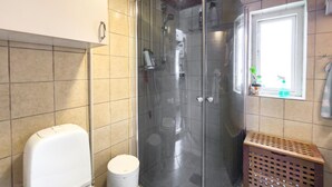 Ein Badezimmer mit Duschkabine, Toilette, Mülleimer und einem Fenster mit Pflanze.