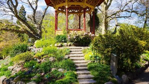 Ein Gartenpavillon mit roter und gelber Struktur, umgeben von einem Garten mit verschiedenen Pflanzen und einer Steintreppe, die hinauf führt.