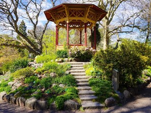 Ein Gartenpavillon mit roter und gelber Struktur, umgeben von einem Garten mit verschiedenen Pflanzen und einer Steintreppe, die hinauf führt.