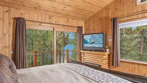 Ein Schlafzimmer einer Holzhütte mit Bett, Fernseher und Fenster mit Blick auf Bäume.