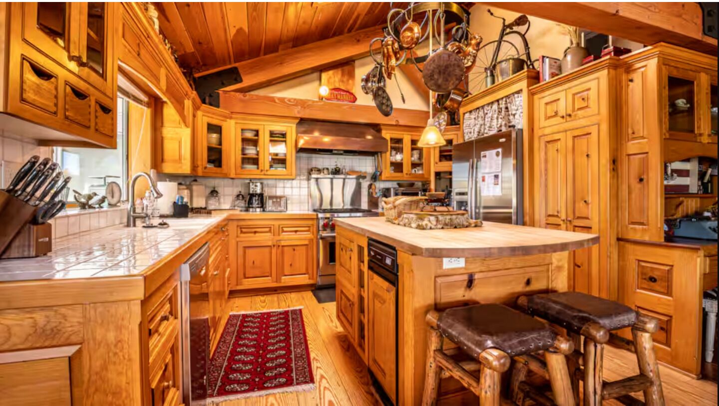 Una cocina con armarios de madera, una isla central, taburetes de bar y utensilios de cocina colgantes.
