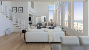 Un amplio salón con un sofá blanco, una escalera y grandes ventanales con vistas a la playa.
