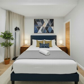 Ett välordnat hotellrum med en stor säng, två sängbord, en golvlampa, en krukväxt och ett geometriskt konstverk på väggen.