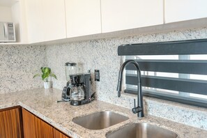 Una cocina con encimeras de granito, cafetera, licuadora, pileta doble y ventana con persianas.