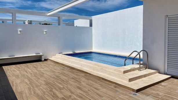 Een modern buitenzwembad met een houten terras, een witte muur en een heldere lucht.