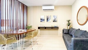 Een moderne woonkamer met een eethoek, een grijze bank, een houten eettafel, rieten stoelen, een wandairco en lijstwerken.