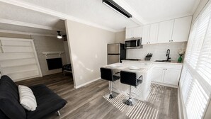 A modern kitchen with white cabinets, a microwave, and a refrigerator. There is a black sofa with a white pillow in a living area.