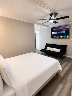 A bedroom with a bed, a black sofa, a ceiling fan, and a TV displaying a home screen.