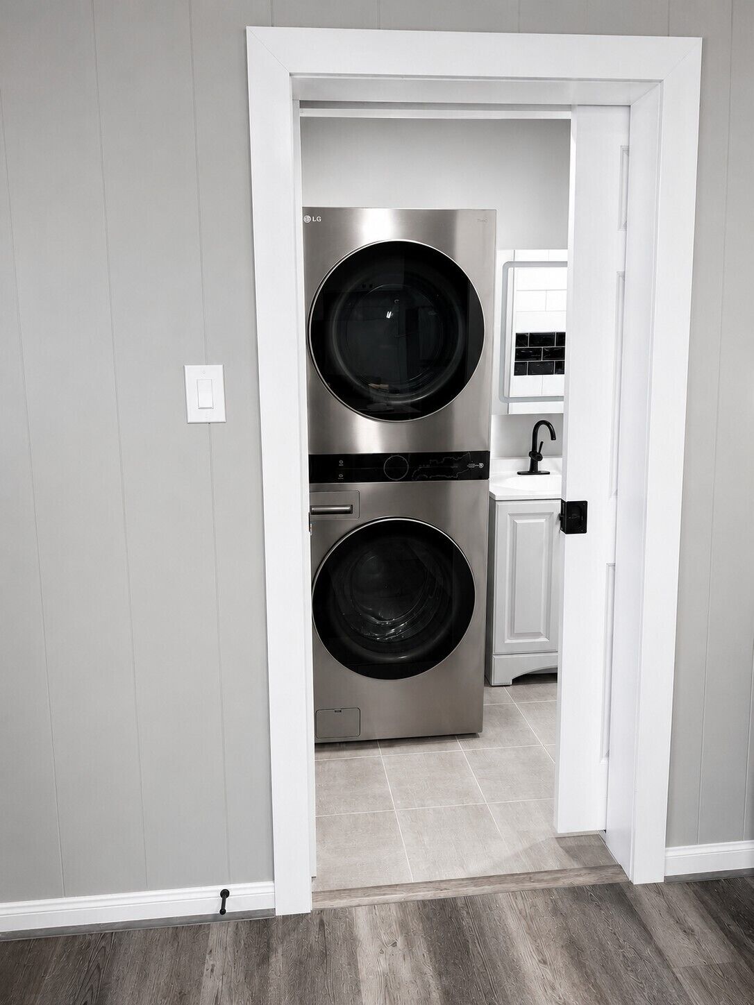 A laundry room with two stacked front-loading washing machines, a white sink, and a door leading to another room.