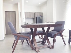 Ein moderner Essbereich mit einem zentralen Tisch und vier Stühlen, im Hintergrund eine Küche und eine Tür links.