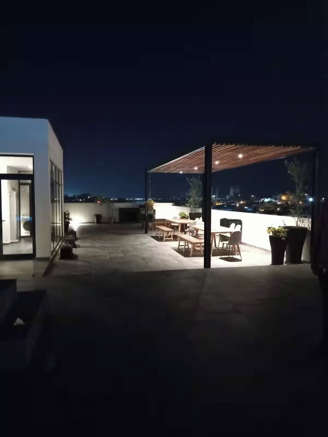 A rooftop patio with outdoor seating, a pergola, and a view of the cityscape at night.