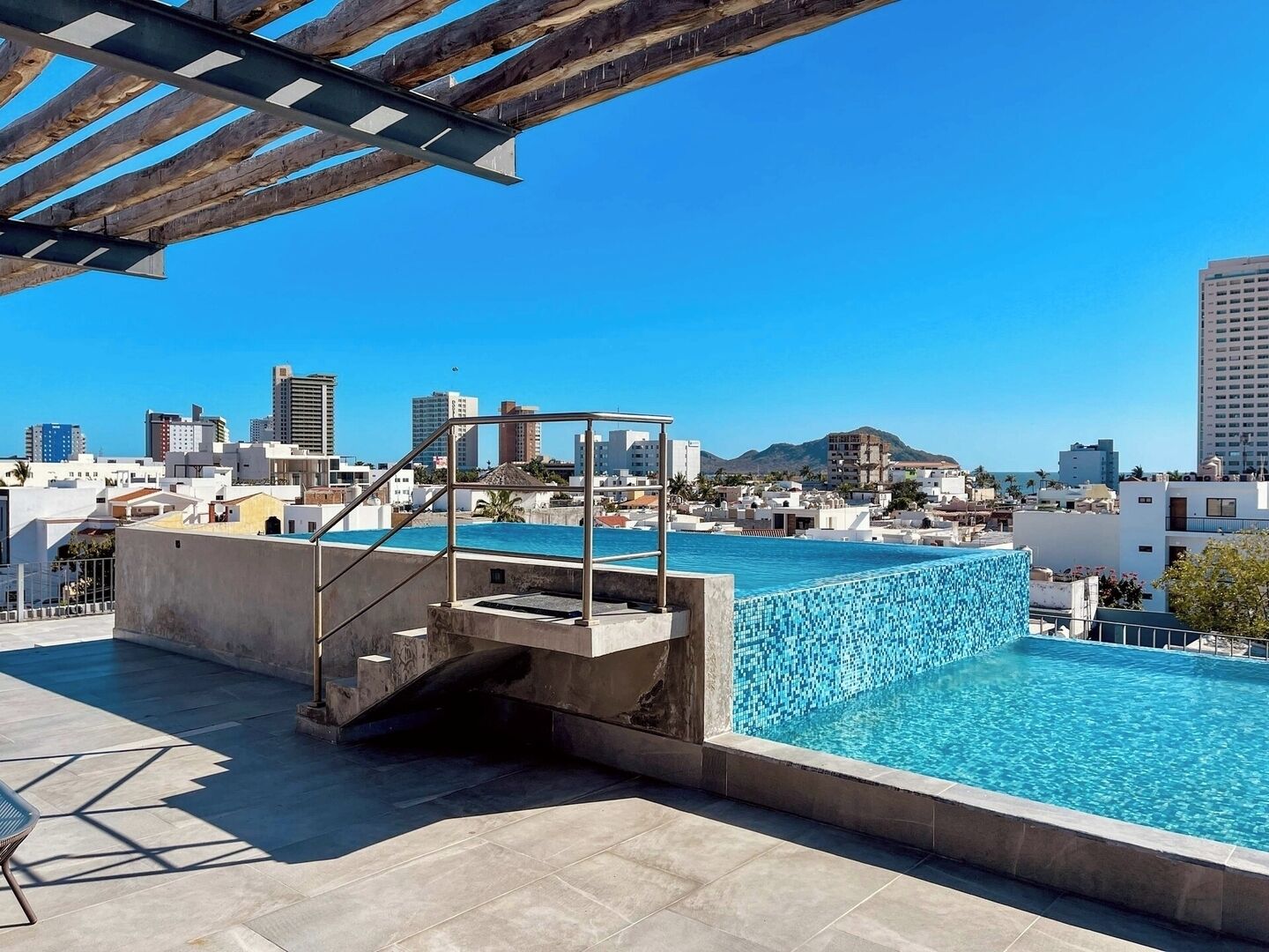 Una piscina al terrat amb vista a la ciutat, un plataforma de busos i un cel blau clar.