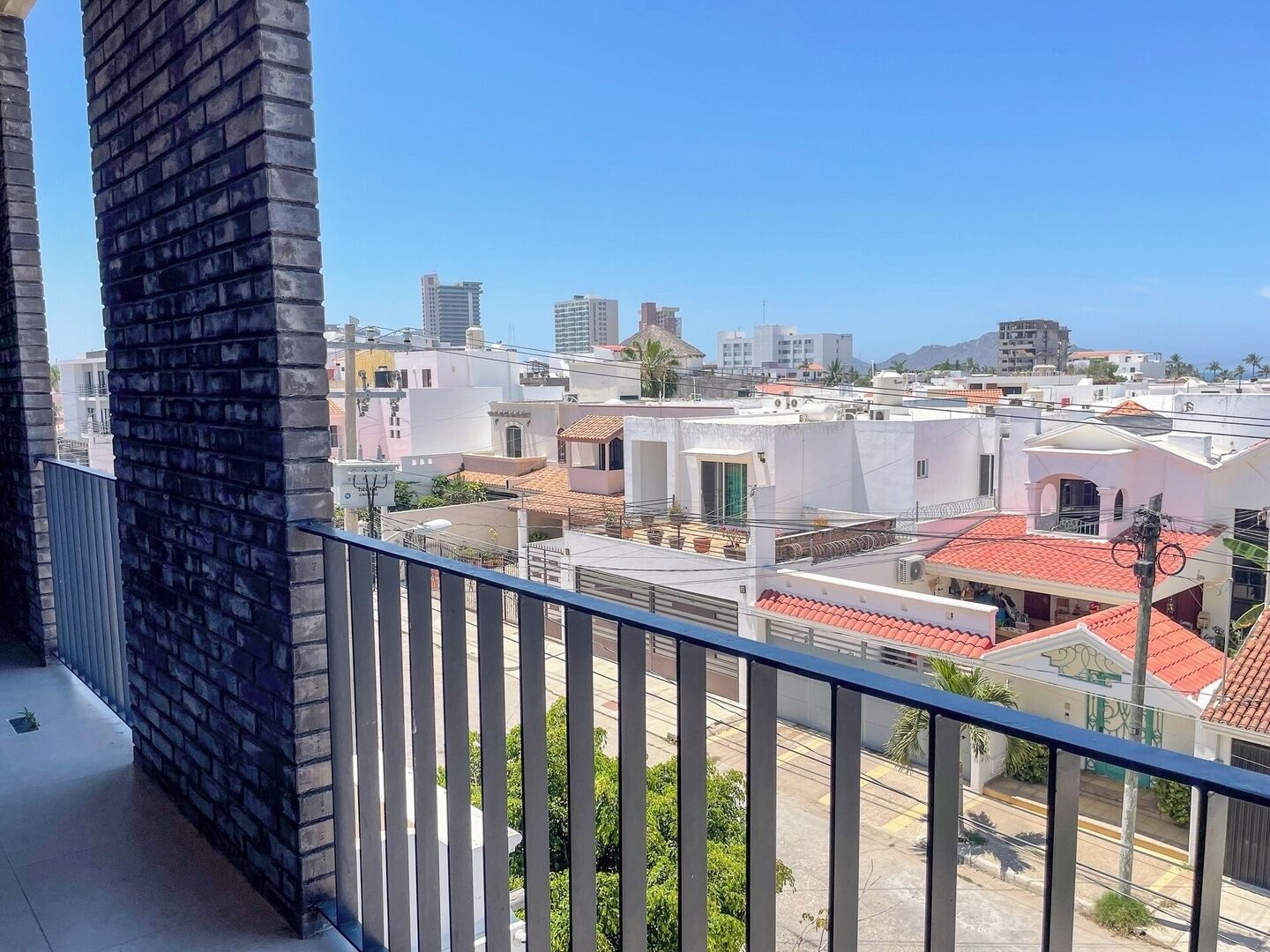 Uma varanda com vista para uma área residencial, incluindo edifícios com telhados de terracota e um céu azul límpido.