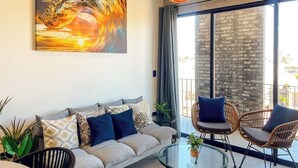 A modern living room with a sofa, two wicker chairs, a glass coffee table, and a large painting of a wave. - Charming Apartment w/ Pool 3 Blocks to Beach (Mazatlan)