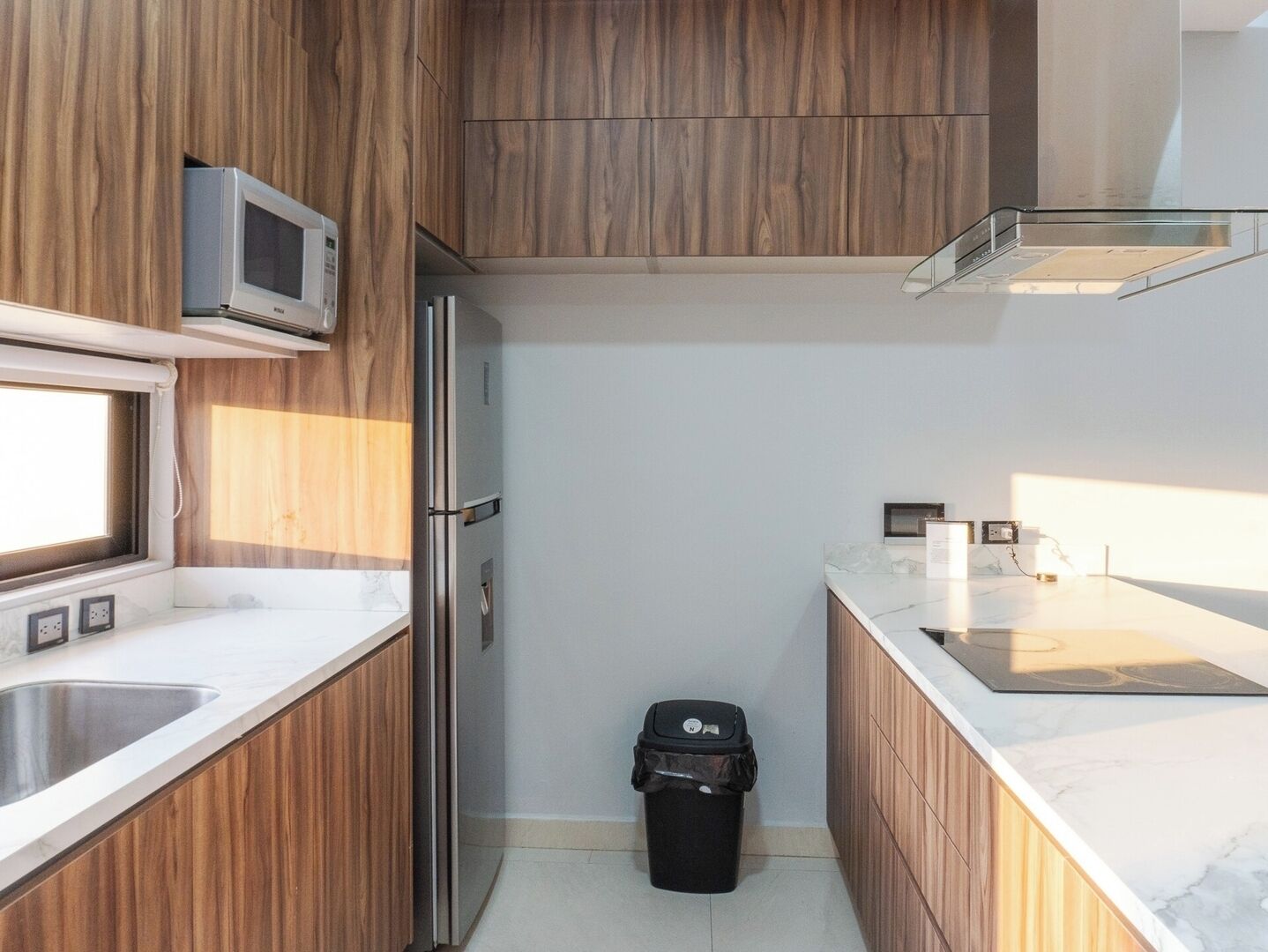 Una cocina compacta con armarios de madera, un refrigerador de acero inoxidable, un microondas y un cubo de basura.